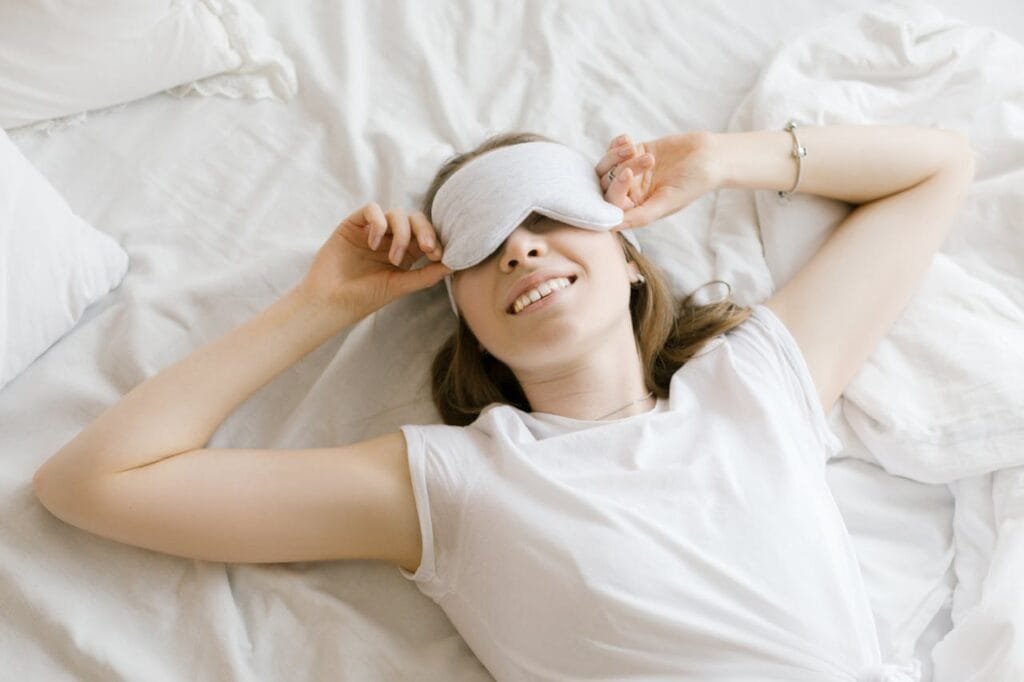 Woman smiling while wearing a sleep mask in a bright white bedroom